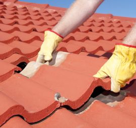 Roof repairs, worker with yellow gloves replacing red tiles or shingles on house with blue sky as background and copy space.